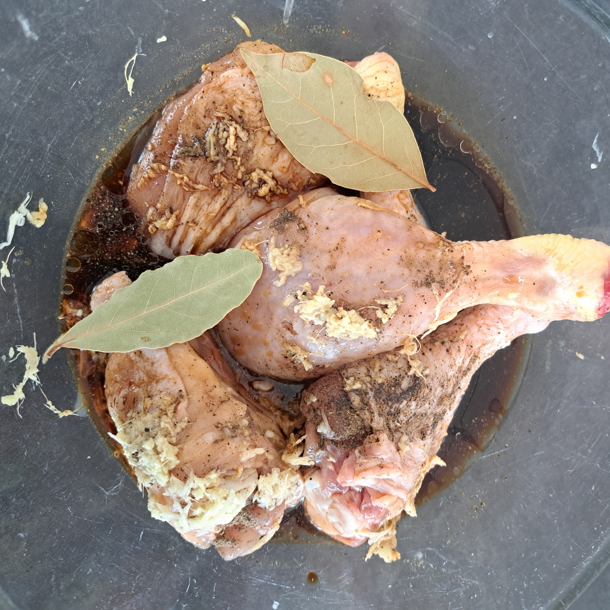 Hähnchenstücke in einer durchsichtigen Glasschüssel mit Marinade aus Sojasauce, Essig, Ingwer, Knoblauch, Lorbeerblättern und Pfeffer, bereit zum Marinieren.