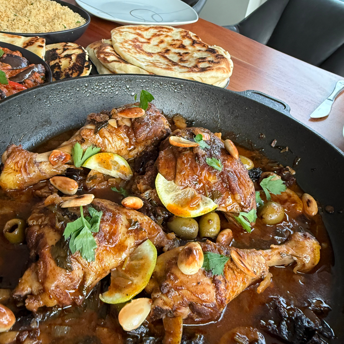 Frisch gekochtes marokkanisches H&auml;hnchen mit Oliven, Zitronen, Pflaumen und Ras el Hanout in der Pfanne, auf einem Holztisch garniert mit frischer Petersilie, Zitronenscheiben und gebr&auml;unten Mandeln. Daneben Beh&auml;lter mit Couscous, Fladenbrot und sudanesischem Auberginensalat.
