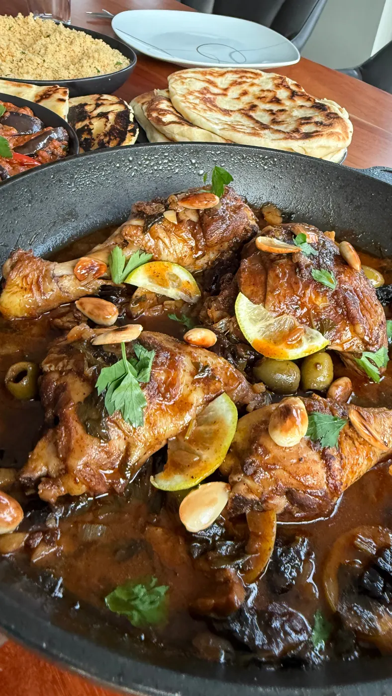 Frisch gekochtes marokkanisches Hähnchen mit Oliven, Zitronen, Pflaumen und Ras el Hanout in der Pfanne, auf einem Holztisch garniert mit frischer Petersilie, Zitronenscheiben und gebräunten Mandeln. Daneben Behälter mit Couscous, Fladenbrot und sudanesischem Auberginensalat.