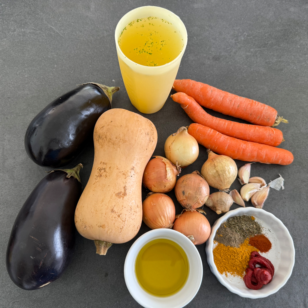 Frische Zutaten für einen afrikanischen Gemüseeintopf, darunter gewürfelter Butternut-Kürbis, Auberginen, Karotten, Zwiebeln, Knoblauch, Ingwer, Currygewürz, Cayennepfeffer, Thymian und Gemüsebrühe, appetitlich auf der Küchenbank arrangiert.