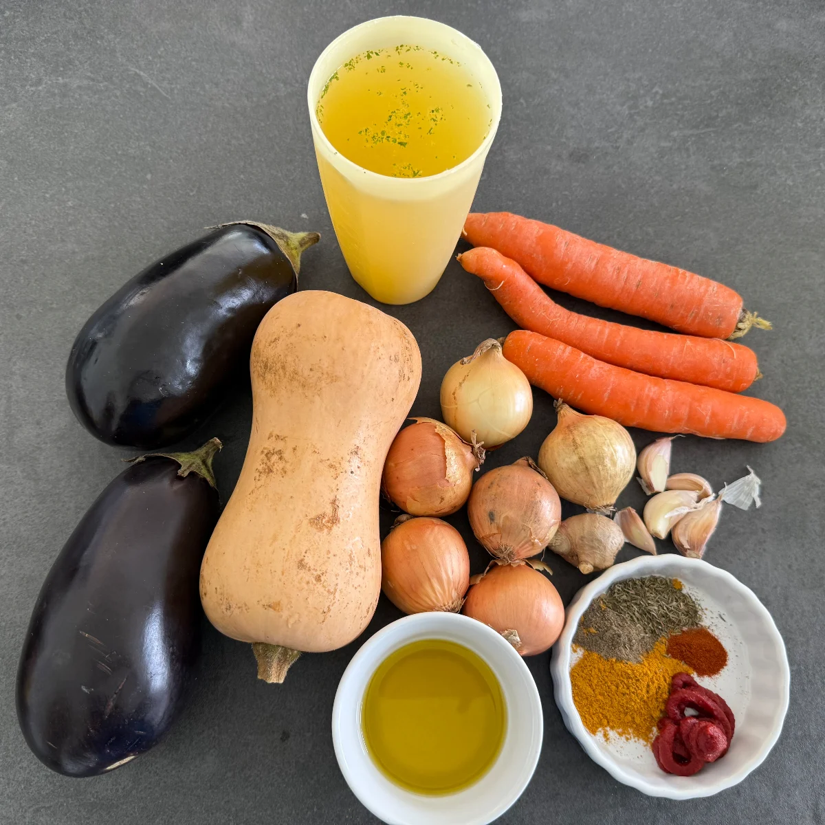 Frische Zutaten für einen afrikanischen Gemüseeintopf, darunter gewürfelter Butternut-Kürbis, Auberginen, Karotten, Zwiebeln, Knoblauch, Ingwer, Currygewürz, Cayennepfeffer, Thymian und Gemüsebrühe, appetitlich auf der Küchenbank arrangiert.