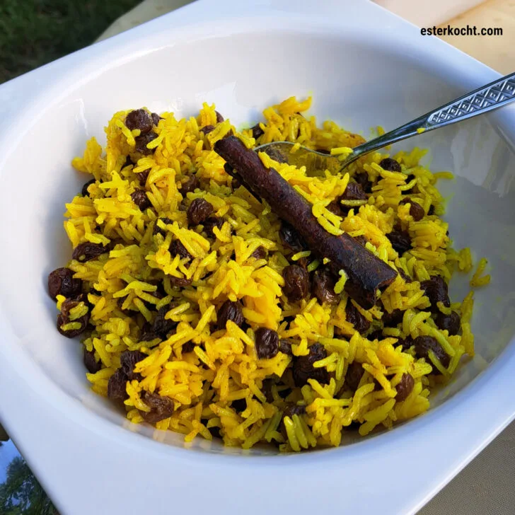 A bowl of fluffy yellow rice with raisins, topped with a cinnamon stick.