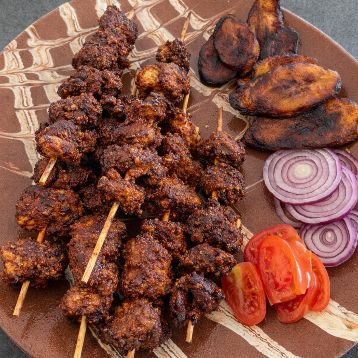Gegrilltes Suya Chicken auf einem Teller mit Tomatenscheiben, roten Zwiebeln und gebratenen Kochbananen